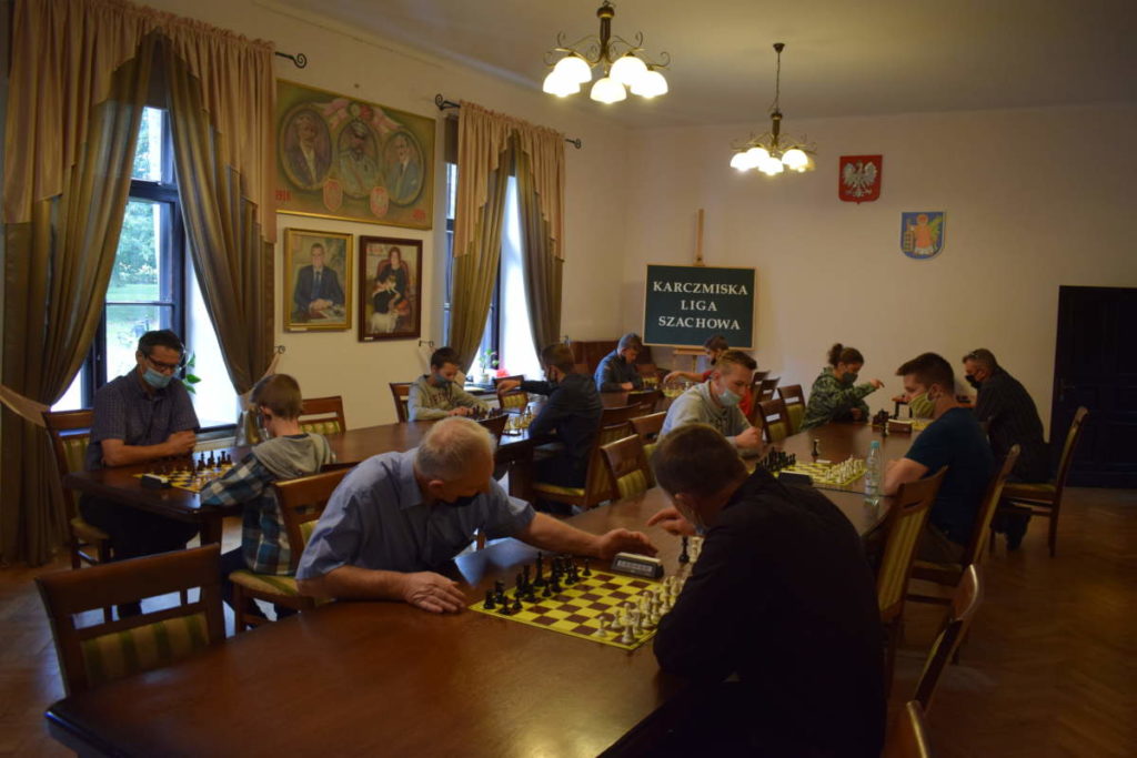 W sali ustawione są dwa rzędy stołów. Przy nich na pierwszym planie dwóch mężczyzn rozgrywa partię szachów. W tle grają pozostali uczestnicy.