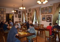 Na pierwszym planie przy stole dwóch uczestników turnieju rozgrywa partię szachów. W tle grają pozostali uczestnicy.