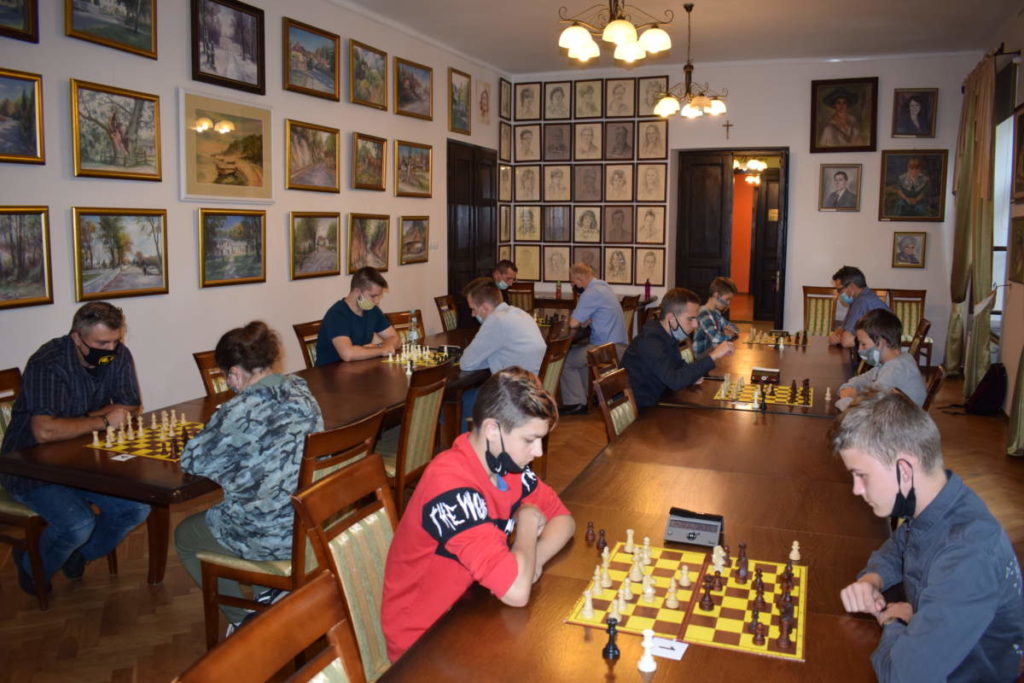 Szachiści grają w szachy. Na pierwszym planie widoczny jeden z uczestników oraz plansza do gry. W tle grają pozostali uczestnicy.