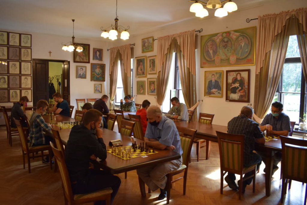 Na pierwszym planie przy stole dwóch uczestników turnieju rozgrywa partię szachów. W tle grają pozostali uczestnicy.