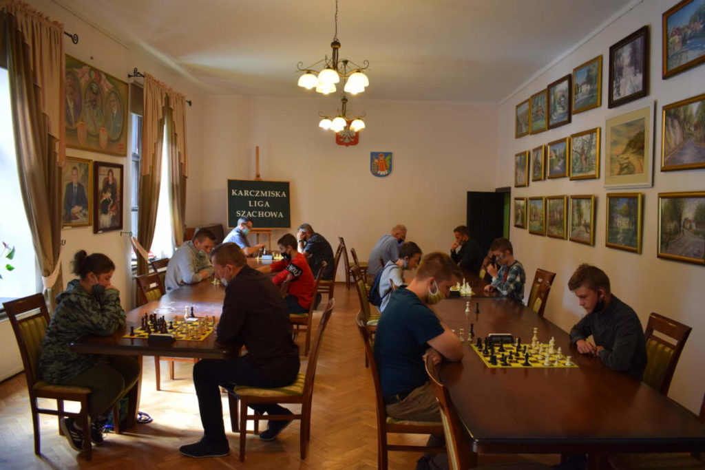 Na zdjęciu widoczne dwa rzędy stołów ustawione prostopadle do siebie. Przez środek jest przez środek jest przejście. Przy stołach uczestnicy turnieju grają w szachy.