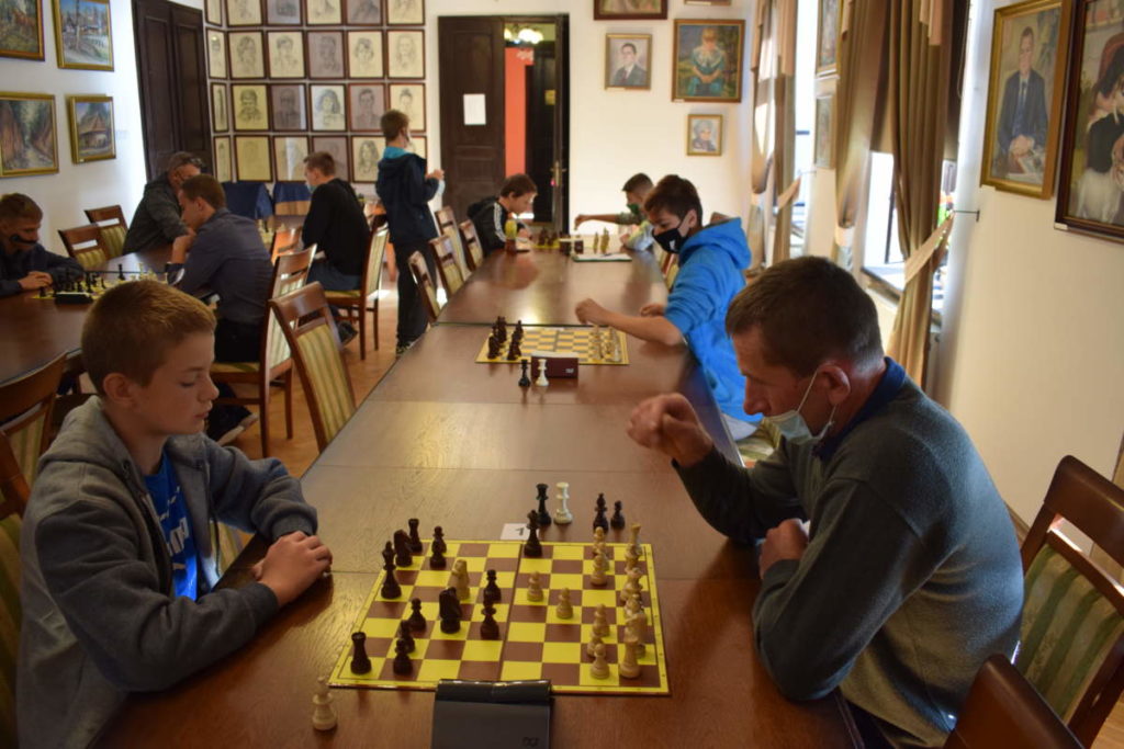 W sali ustawione są dwa rzędy stołów. Przy nich zawodnicy siedzą i grają w szachy. Jeden z uczestników stoi przy stole.