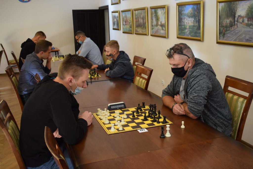 Przy stołach widać trzy pary uczestników. Zawodnicy w maseczkach albo półprzyłbicach grają w szachy.
