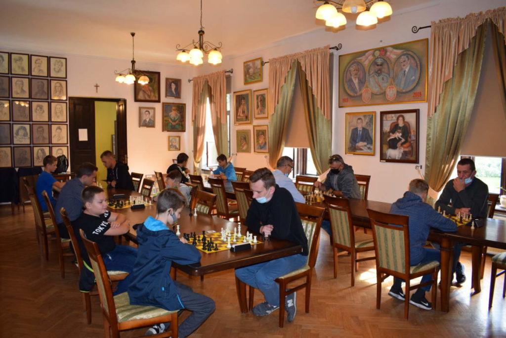 W sali przy stołach grają szachiści. Na ścianach wiszą obrazy.