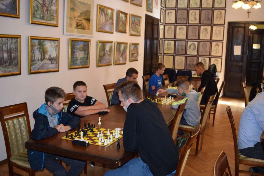 Zbliżenie na jeden stół. Na pierwszym planie dwóch uczestników rozgrywa partię szachów. Obok jeden z zawodników przygląda się grze. W tle widoczni pozostali gracze.