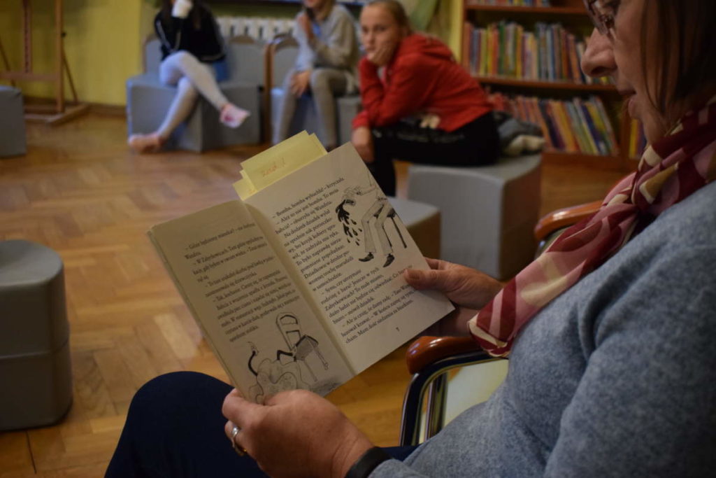 Zabawy integracyjne. Na pierwszym planie siedząca kobieta czyta książkę. W tle widoczne dzieci siedzące na pufach. Za nimi pułki z książkami.