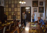 Na zdjęciu widoczna jest cała sala. Przy stołach ustawionych w dwóch rzędach siedzą grający w szachy zawodnicy. Jedna osoba stoi przy drzwiach.