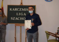 Zdjęcie zawodnika turnieju trzymającego w ręku dyplom. Mężczyzna stoi na tle tablicy ustawionej na sztaludze z napisem „KARCZMISKA LIGA SZACHOWA”.