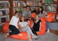 Zdjęcie grupowe. Kilka osób siedzi na dużych pomarańczowych pufach. Patrzą przed siebie – pozują do zdjęcia.