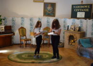 Dwie młode dziewczyny stoją obok siebie. Trzymają kartki z tekstem i czytają jego fragmenty. Za plecami jest dekoracja.