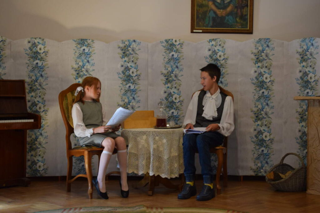 Przy niewielkim stole siedzi na krzesłach dwoje młodych ludzi. Czytają trzymane kartki z tekstem. Siedzą na tle sztucznej ściany obitej tapetą. Z jednej strony widoczna część pianina, a z drugiej fragment kominka.
