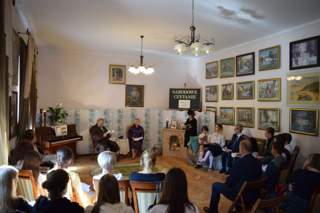 Widok ogólny na całą salę. Na pierwszym planie siedzący na krzesłach uczestnicy wydarzenia. W tle widać dekorację przy której pozostali uczestnicy czytają.