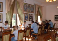 Przy stołach ustawionych przy oknach siedzą zawodnicy i grają w szachy. Na ścianie wiszą obrazy.