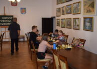 Przy stołach ustawionych przy ścianie zawodnicy grają w szachy. na ścianie wiszą obrazy.