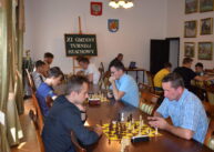 Przy stołach zawodnicy grają w szachy. Za nimi w tle tablica z napisem XI GMINNY TURNIEJ SZACHOWY. Dalej widoczne są otwarte drzwi.