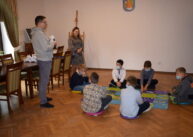 W sali dzieci siedzą na podłodze, dwie dorosłe osoby stoją. Jedna z nich trzyma niewielkiego robota.