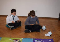 Na zdjęciu dwoje dzieci siedzi na podłodze z tabletem w ręku. Obok są dwa roboty. Za dziećmi pusta ściana.