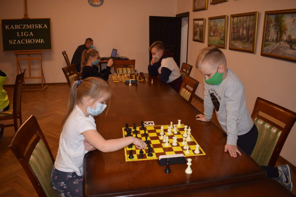 Przy stole para zawodników gra w szachy. W tle widać kolejną parę zawodników turnieju.