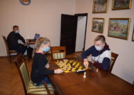 Przy stole para zawodników gra w szachy. W tle widać mężczyznę siedzącego przy stole. Obok otwarte drzwi.