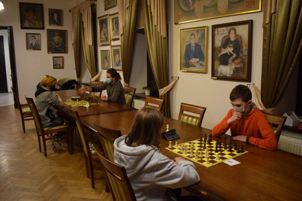 Przy stole para zawodników gra w szachy. Z tyłu na ścianie wiszą obrazy.