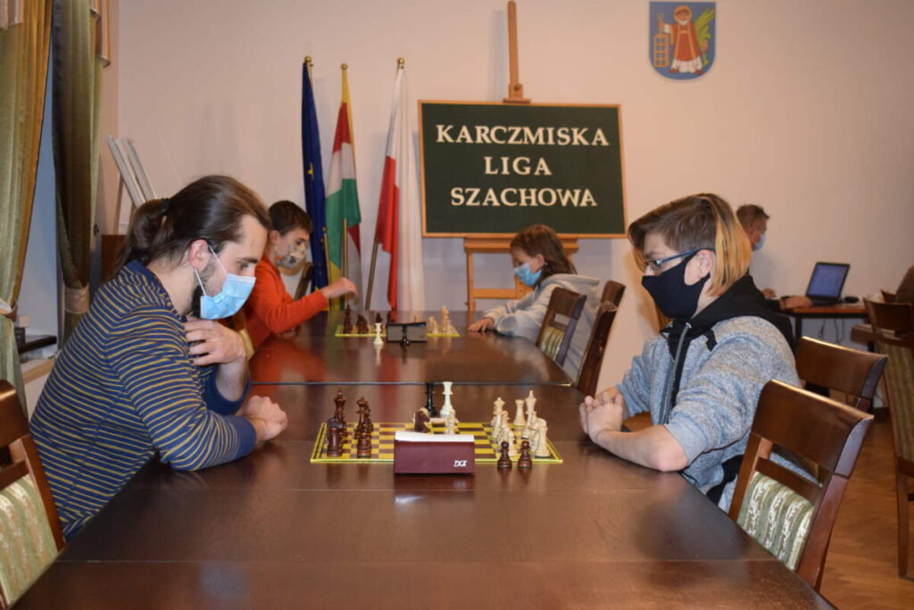 Widok na rząd stołów, przy których grają zawodnicy. W tle widoczna zielona tablica z napisem KARCZMISKA LIGA SZACHOWA. Obok niej trzy flagi.