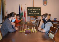 Widok na rząd stołów, przy których grają zawodnicy. W tle widoczna zielona tablica z napisem KARCZMISKA LIGA SZACHOWA. Obok niej trzy flagi.