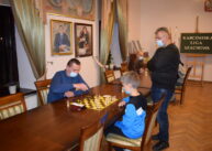 Przy stole para zawodników gra w szachy. Obok jednego z uczestników stoi mężczyzna.