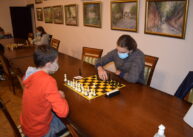 Przy stole para zawodników gra w szachy. Z tyłu na ścianie wiszą obrazy.