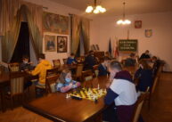 Przy stole para zawodników gra w szachy. W tle widać pozostałych uczestników turnieju.