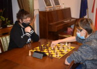 Przy stole para zawodników gra w szachy. W tle widać pianino.