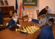 Przy stole para zawodników gra w szachy. W tle widoczna zielona tablica z napisem KARCZMISKA LIGA SZACHOWA. Obok niej trzy flagi.