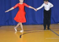 Na zdjęciu widoczna para tańcząca para dzieci. Dziewczynka jest w czerwonej sukience, chłopiec w czarnych spodniach i białej bluzce.