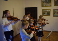 Grupa dzieci gra na skrzypcach. W tle widoczne drzwi i wiszące obrazy na ścianach.