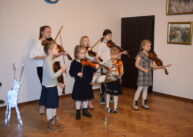 Grupa dzieci gra na skrzypcach. W tle widoczne drzwi i wiszące obrazy na ścianach.