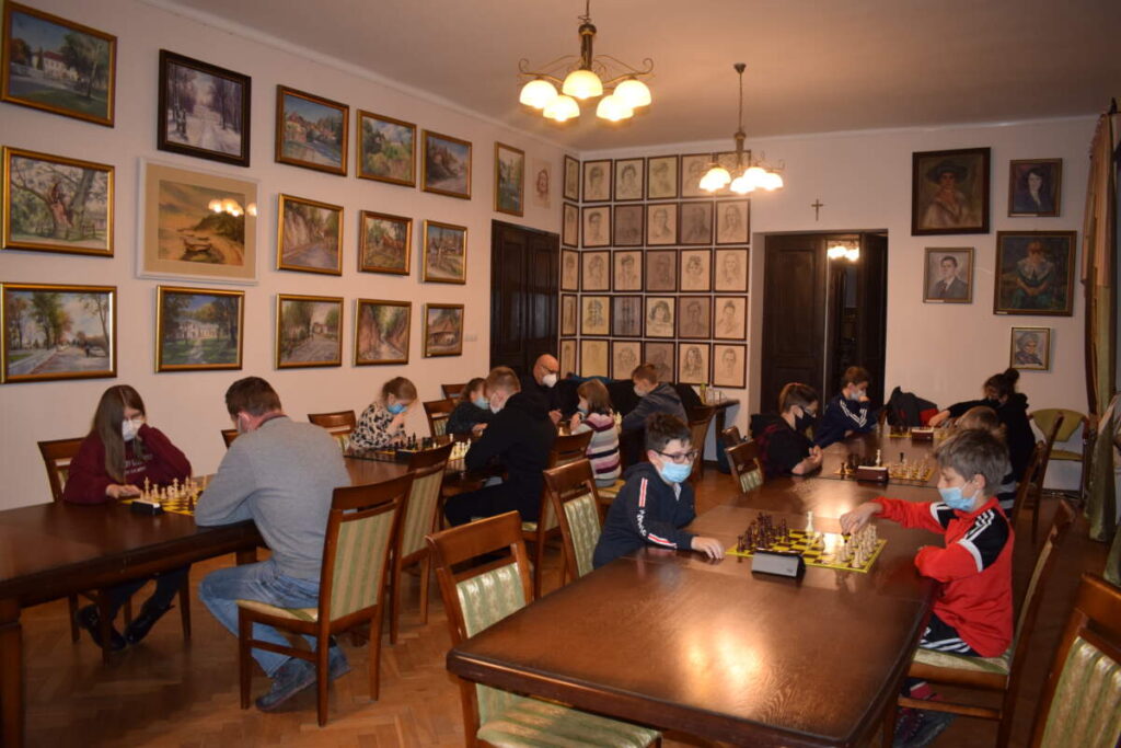 Widok na całą salę. Widać dwa rzędy stołów, przy których uczestnicy turnieju grają w szachy.