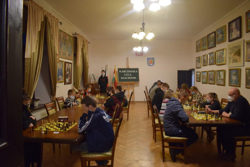 Widok na całą salę. Przy ustawionych równolegle do siebie stołach siedzą zawodnicy i grają w szachy.