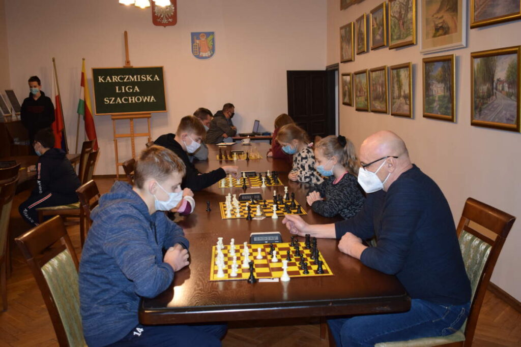 Przy rzędzie stołów ustawionych przy ścianie zawodnicy grają w szachy. W tle widoczna tablica z napisem KARCZMISKA LIGA SZACHOWA.
