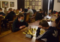 Przy stole para zawodników gra w szachy. W tle widać pozostałych uczestników turnieju.