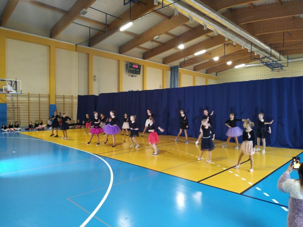 Widok na całą halę sportową. Grupa dziewczynek w kolorowych sukienkach i czarnych bluzkach tańczy na tle granatowej kotary.