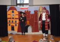 Dwoje aktorów ubranych w historyczne stroje stoi na tle dekoracji. Dekorację stanowi malowidło zamku.
