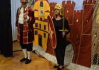 Dwoje aktorów ubranych w historyczne stroje stoi na tle dekoracji. Jeden trzyma tarczę i miecz. Dekorację stanowi malowidło zamku.