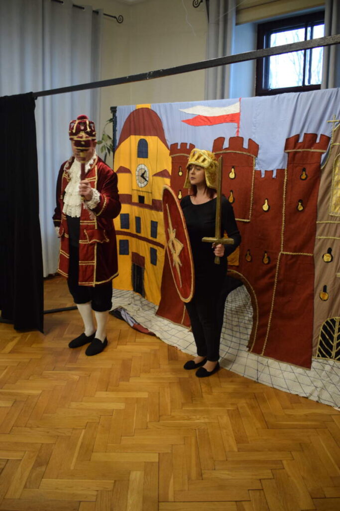 Dwoje aktorów ubranych w historyczne stroje stoi na tle dekoracji. Jeden trzyma tarczę i miecz. Dekorację stanowi malowidło zamku.