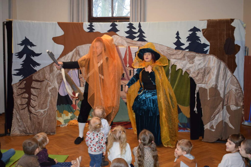 Aktorzy ubrani w kolorowe stroje stoją przed grupą dzieci.