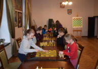 Przy stołach ustawionych przy oknach siedzą zawodnicy i grają w szachy. W tle widać dużą szachownicę na sztaludze.