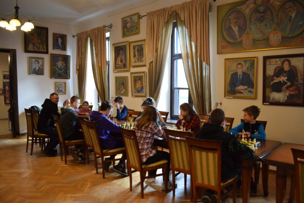 Przy rzędzie stołów ustawionych przy oknach siedzą zawodnicy i grają w szachy.