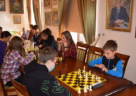 Przy stołach ustawionych przy oknach siedzą zawodnicy i grają w szachy. Na ścianach wiszą obrazy.