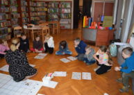 Na podłodze siedzi grupa dzieci. Każdy ma przed sobą kartki z zadaniami. W tle widać stoły, biurko i półki z książkami.