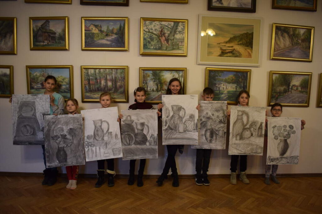 Dzieci stoją na tle ściany z obrazami i prezentują, wykonane na dużych kartonach, swoje prace.