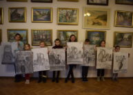 Dzieci stoją na tle ściany z obrazami i prezentują, wykonane na dużych kartonach, swoje prace.