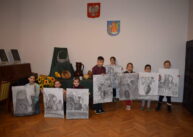 Dzieci na tle kompozycji z wazonów i kwiatów prezentują, wykonane na dużych kartonach, swoje prace.